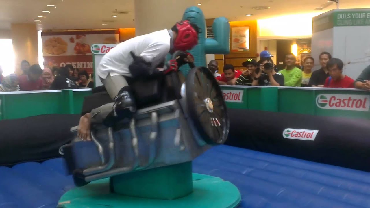 A rider on a Castrol-branded mechanical rodeo machine in a mall, watched by a crowd